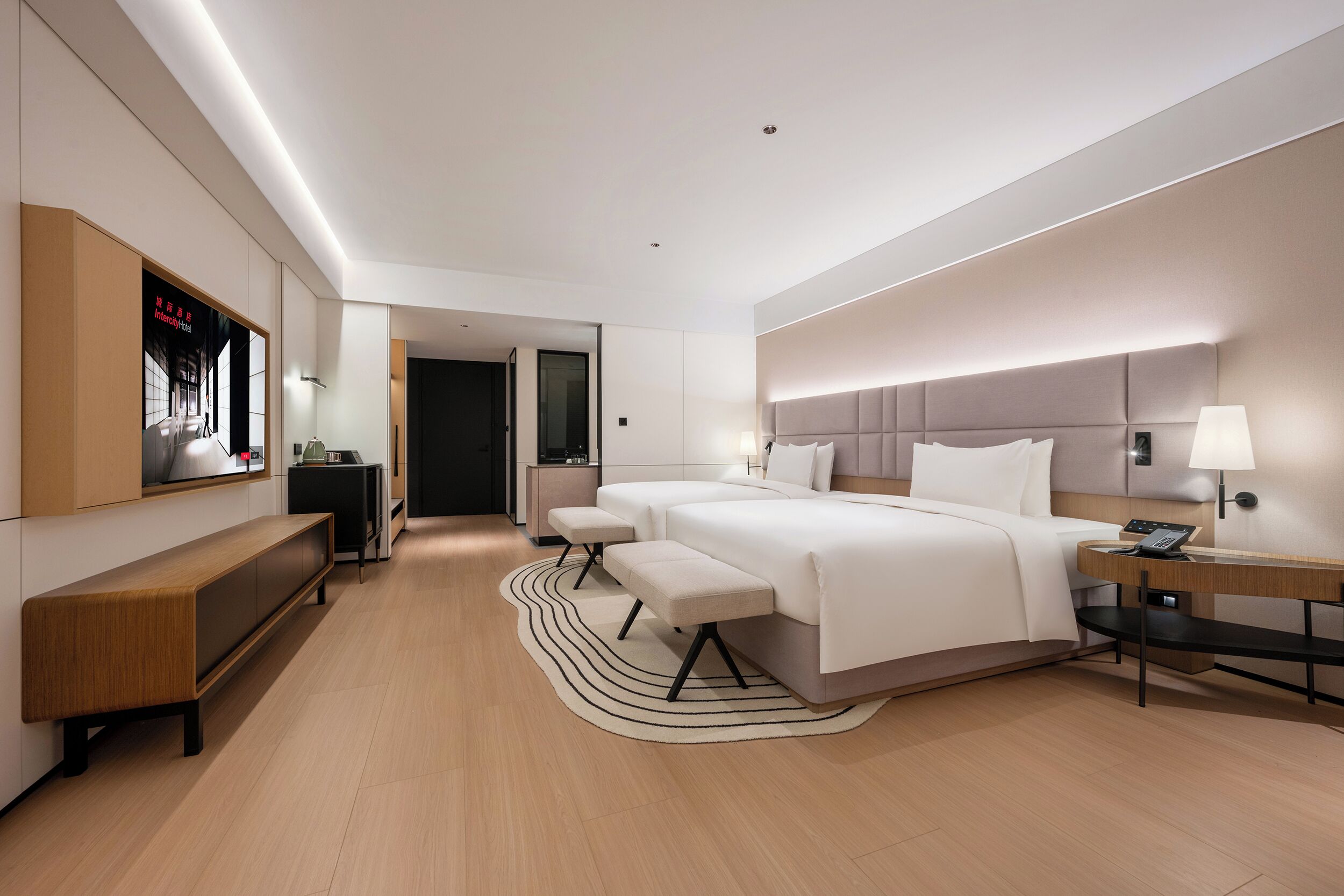 Une chambre d’hôtel moderne dotée d’un grand lit, de tables de chevet, d’un banc, d’un bureau et d’un téléviseur fixé au mur.