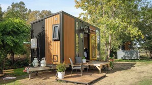 A compact, modern cabin with a wooden exterior, a small deck, a grill, and a seating area with a table and chairs.
