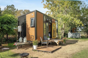 A compact, modern cabin with a wooden exterior, a small deck, a grill, and a seating area with a table and chairs.