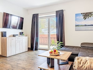 Ein modernes Wohnzimmer mit Holzboden, einer Couch, einem Couchtisch mit Früchten, einem Fernseher und Seeblick durch große Fenster.