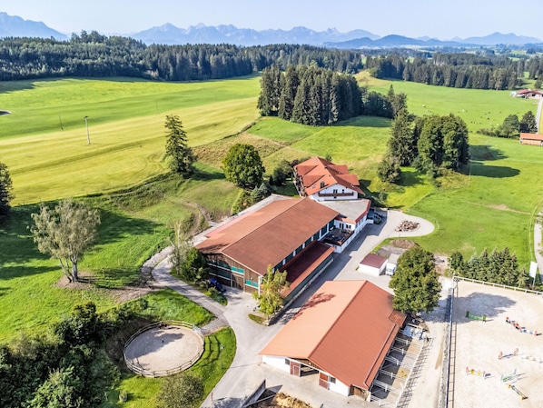An aerial view of a building with a red roof, surrounded by green fields and trees, with mountains in the background.