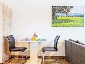 A small, modern room with a wooden wardrobe, a table with two chairs, a laptop, and a painting of a landscape.