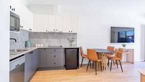 Moderni keittiö, jossa on harmaat kaapit, sisäänrakennettu mikroaaltouuni ja pienikokoinen jääkaappi. Keittiössä on myös ruokailutila, jossa on pyöreä pöytä ja neljä tuolia.