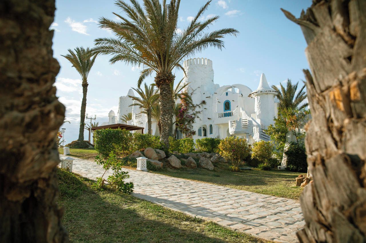 A white building with a stone pathway leading to it, surrounded by palm trees and greenery.