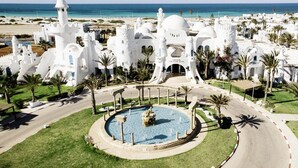 A white building with a fountain in the center, surrounded by palm trees and a beach in the background. - Robinson Djerba Bahiya (Aghir)