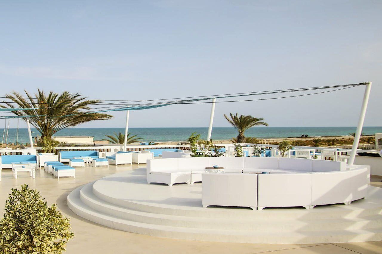 A rooftop lounge with white sofas and tables, palm trees, and a view of the beach.