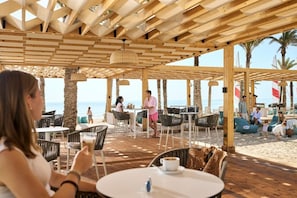 Un café en plein air, avec des tables et des chaises, donnant sur une plage bordée de palmiers et animée par la présence de nombreuses personnes.