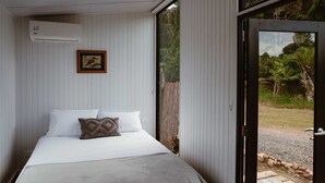 A bedroom with a bed, pillows, a framed picture, an air conditioning unit, and a view through a glass door. - Omoho Stream X by Tiny Away (Kennedy Bay)