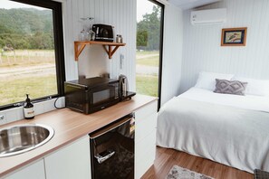 A compact room with a kitchenette, a bed, a window with a view of greenery, and a wall-mounted air conditioner. - Omoho Stream X by Tiny Away (Kennedy Bay)
