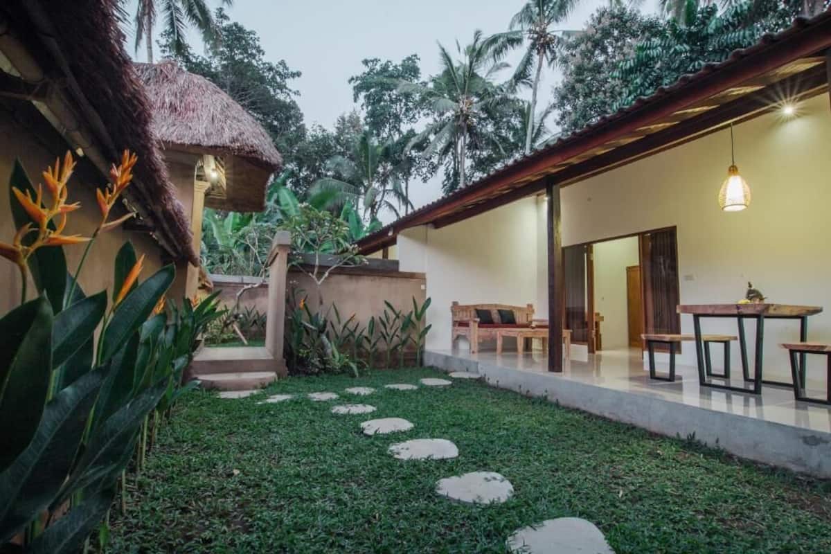 A tropical resort with a thatched-roof structure, a covered patio with outdoor seating, and a well-maintained lawn with stepping stones.