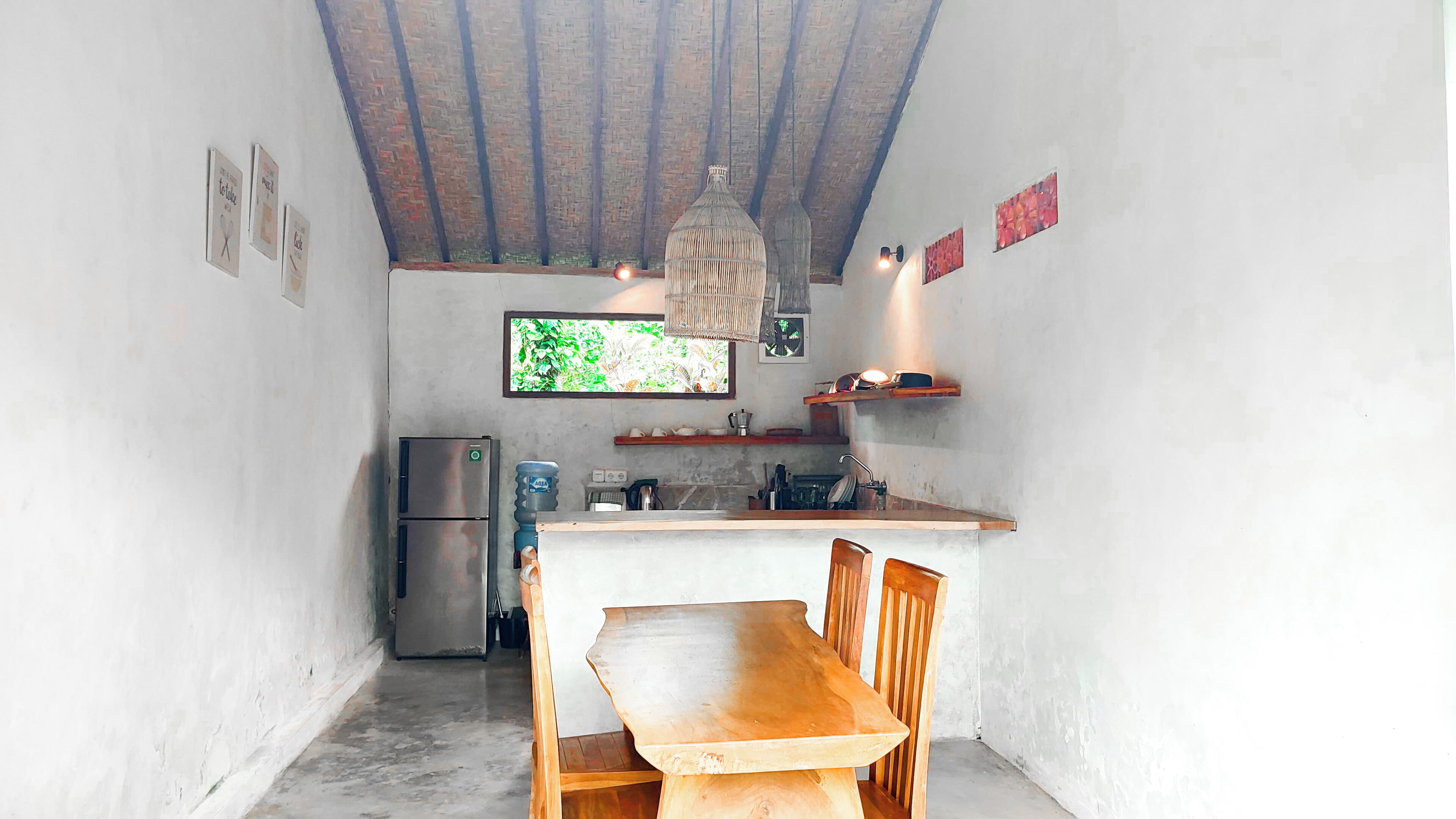 A compact kitchen with a wooden dining table and chairs, a refrigerator, a water dispenser, and a hanging light fixture.
