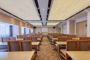 Una sala conferenze con file di tavoli e sedie in legno, pavimento rivestito di moquette e un grande schermo di fronte.