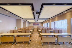 A conference room with rows of tables and chairs, large windows, and a carpeted floor.