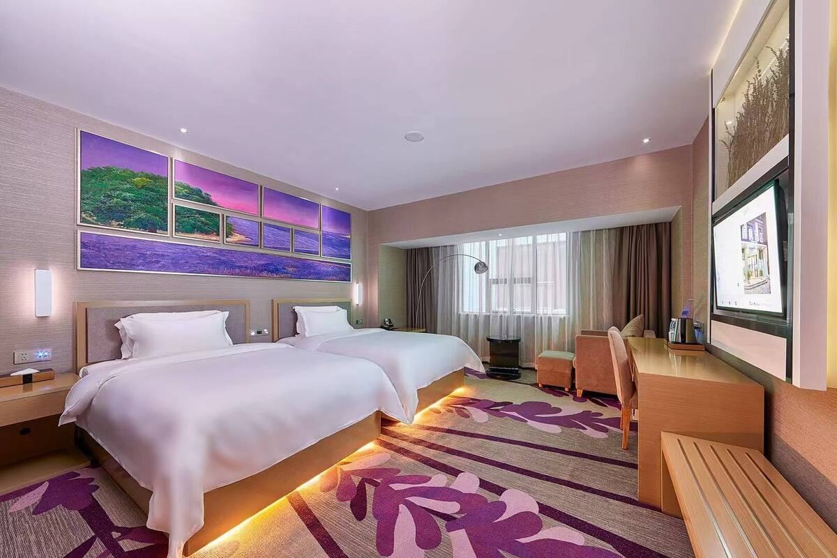 A hotel room with two beds, a desk, a chair, a TV, and a large window with curtains.