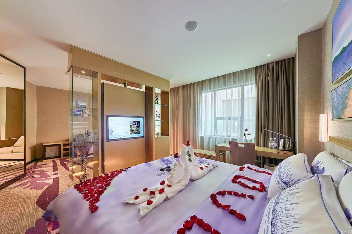 A hotel room with a bed adorned with rose petals and swans, a desk with a chair, a television, and a view of a cityscape through large windows.