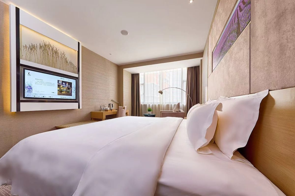 A hotel room with a large bed, a flat-screen TV mounted on the wall, a desk with a lamp, and a window with curtains.