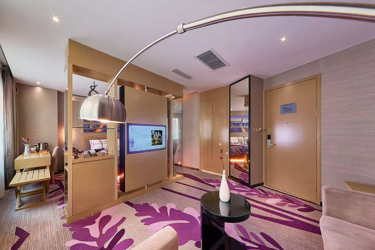 A modern hotel room with a desk, a large mirror, a television, and a comfortable seating area.