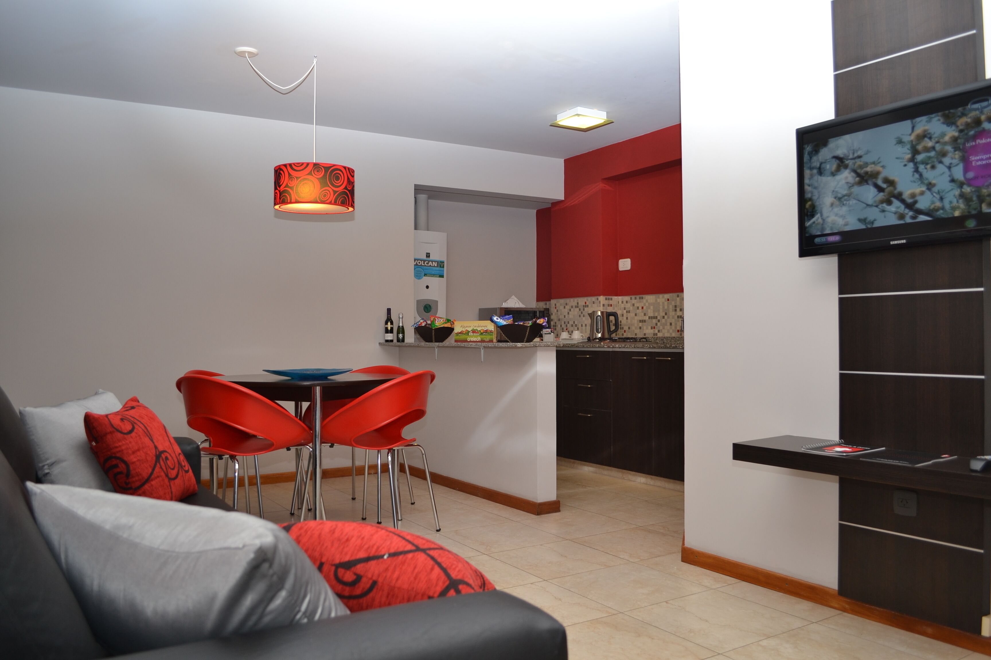 A modern living room with a sofa, red chairs, a TV, and a kitchen area with a red accent wall.