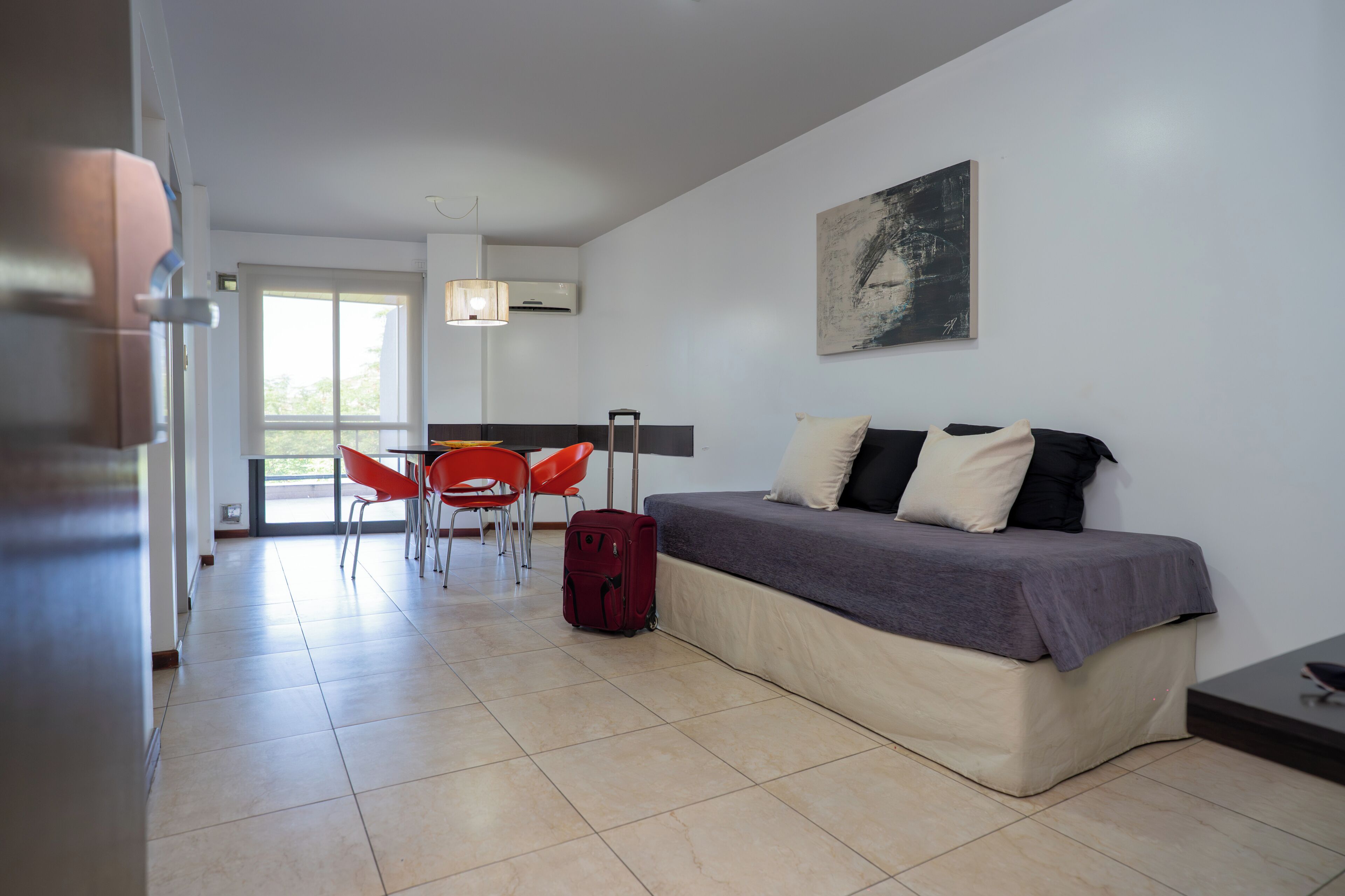 A modern room with a bed, a dining area with red chairs, a red suitcase, and a painting on the wall.