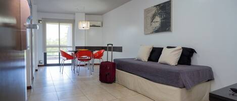 A modern room with a bed, a dining area with red chairs, a red suitcase, and a painting on the wall.