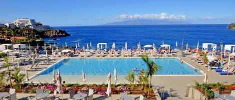 A resort pool area with multiple lounge chairs and umbrellas, a large swimming pool, and a view of the ocean and distant buildings.