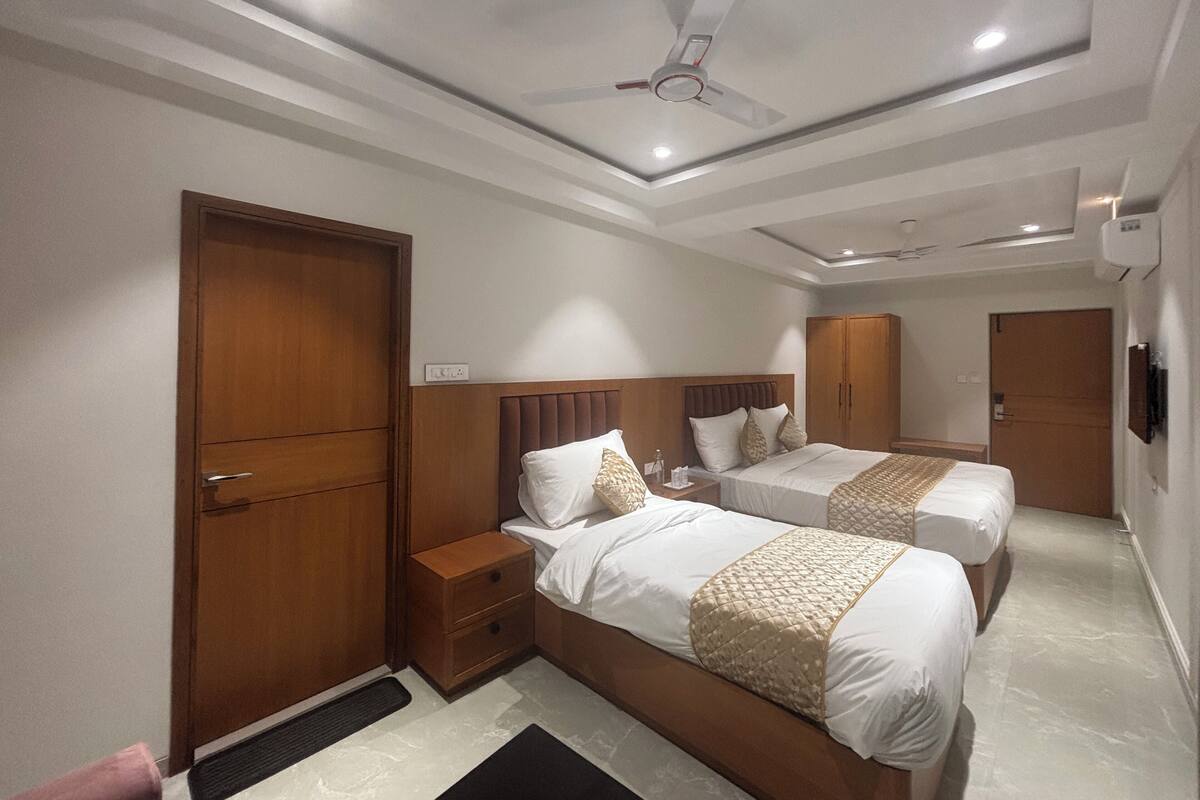 A hotel room with two beds, a wooden wardrobe, a bedside table, a ceiling fan, and an air conditioning unit.