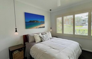A bedroom with a bed, a nightstand, a lamp, a painting, and a window with blinds.