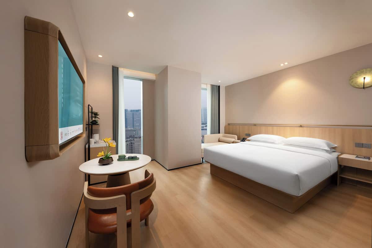 A modern hotel room with a large bed, a desk, and a chair. There is a city view through the window, and the room has a wooden floor and white walls.