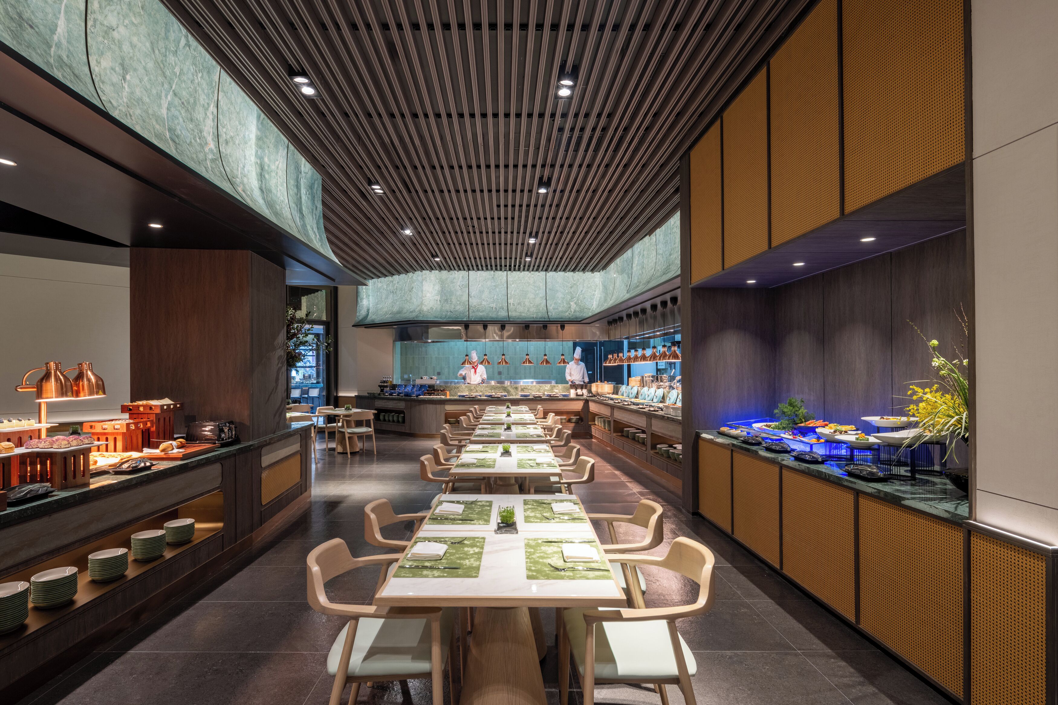 A modern restaurant with long tables, a kitchen in the background, and a wooden ceiling.
