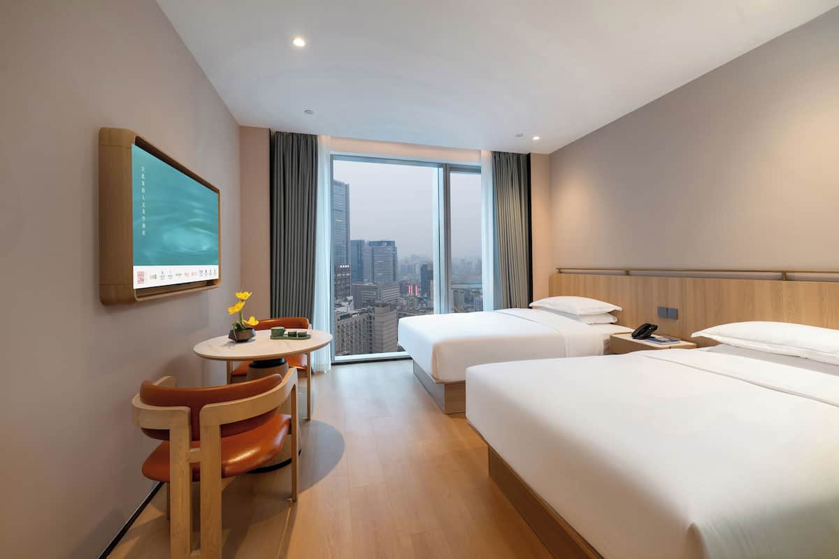 A hotel room with two beds, a desk with a chair, a television mounted on the wall, and a large window offering a city view.