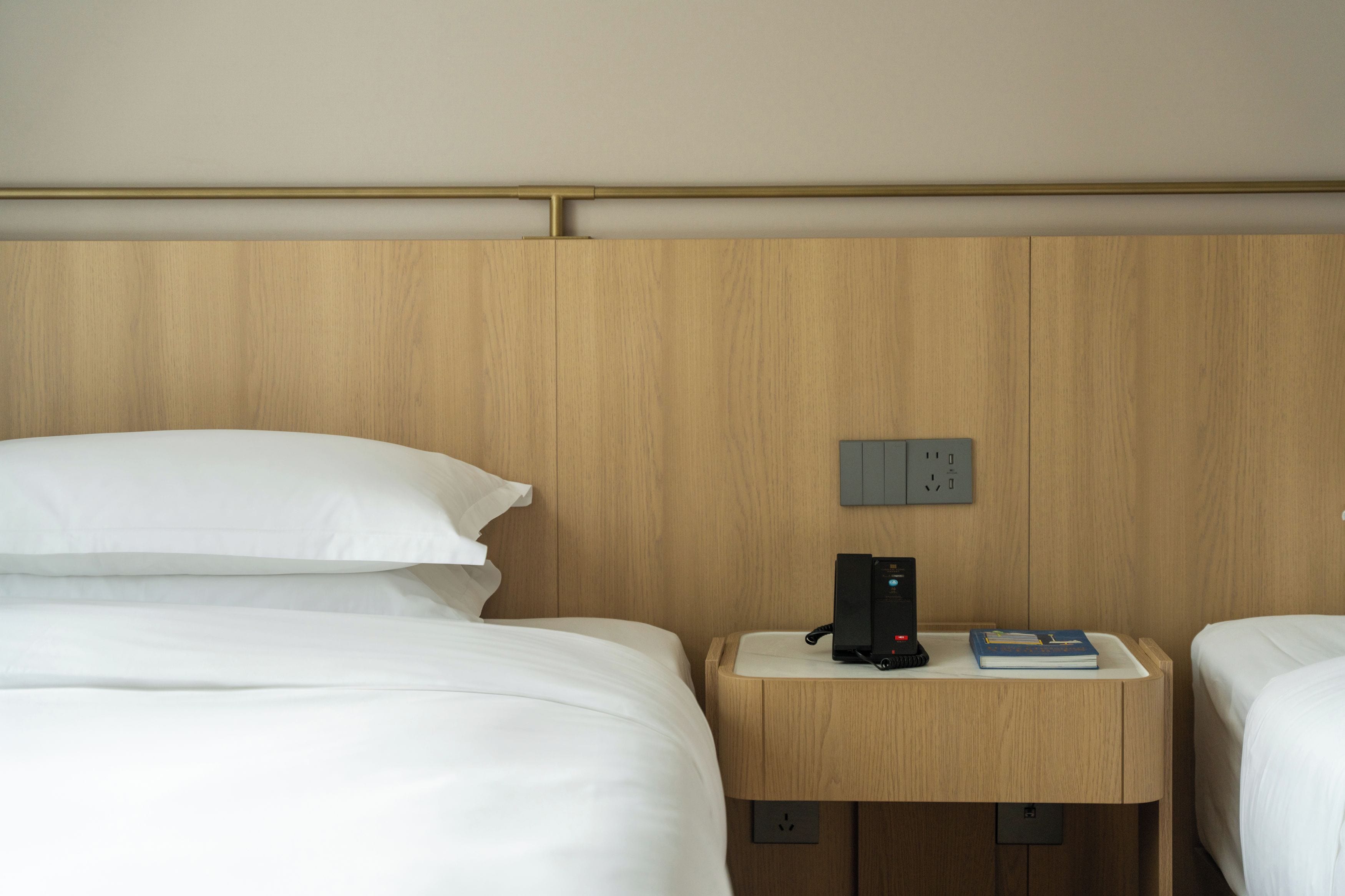 Habitación de hotel con dos camas, cabecera de madera, mesita de noche con un teléfono y un libro, y un enchufe.