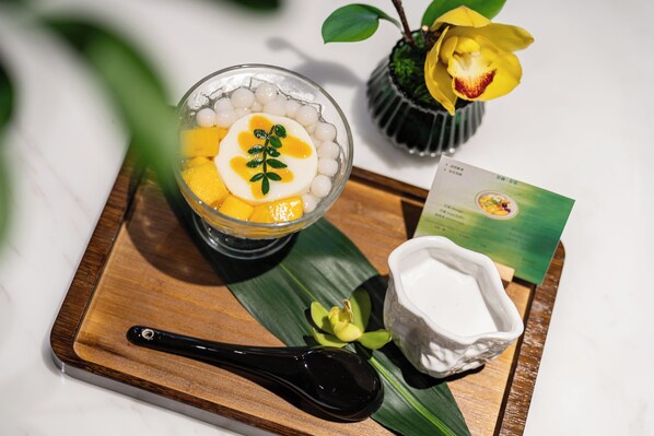 A dessert with mango and white spheres, a spoon, a small white dish, and a card on a wooden tray.