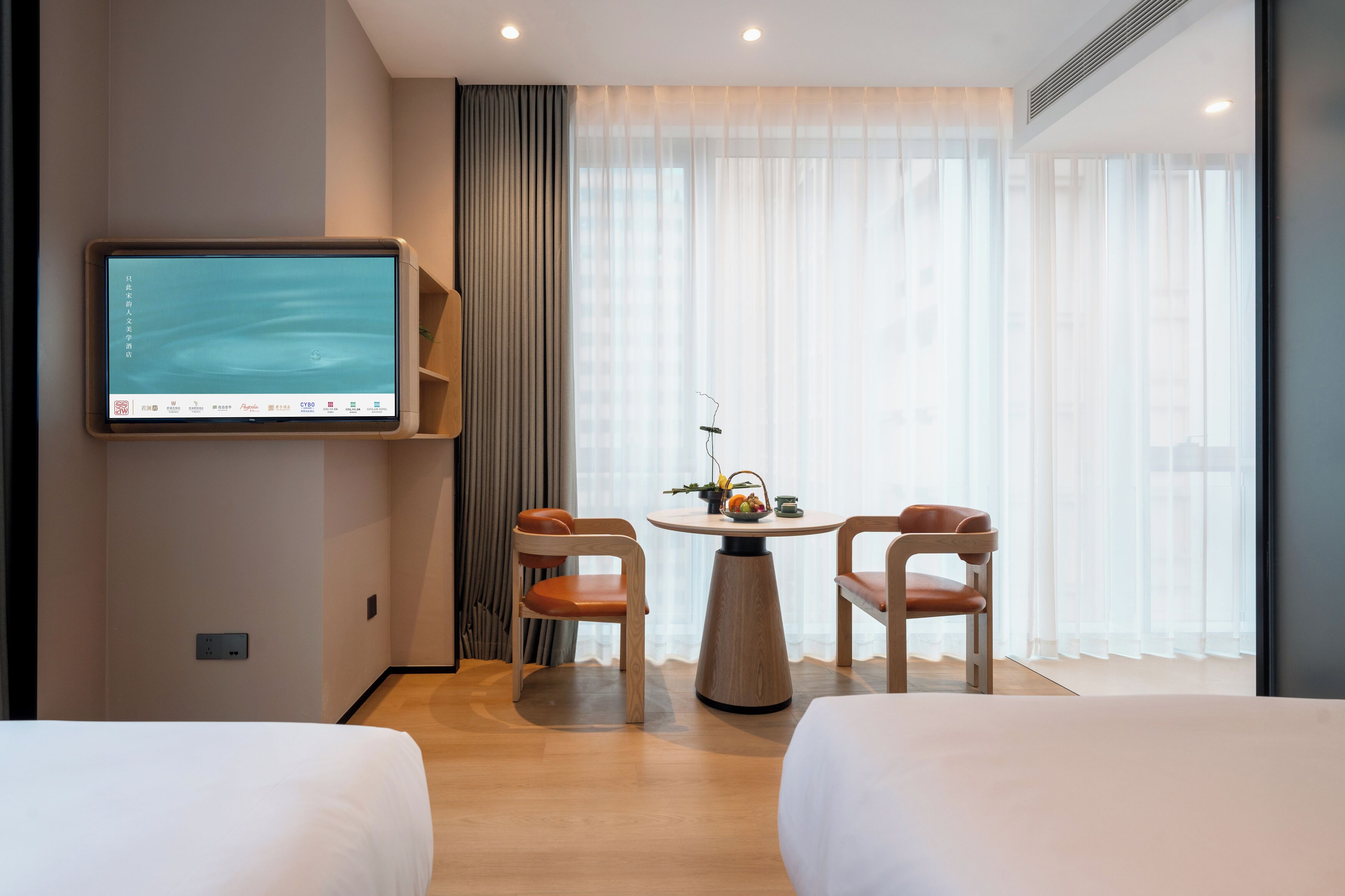 A hotel room with two beds, a TV mounted on the wall, a small round table with chairs, and a large window with sheer curtains.