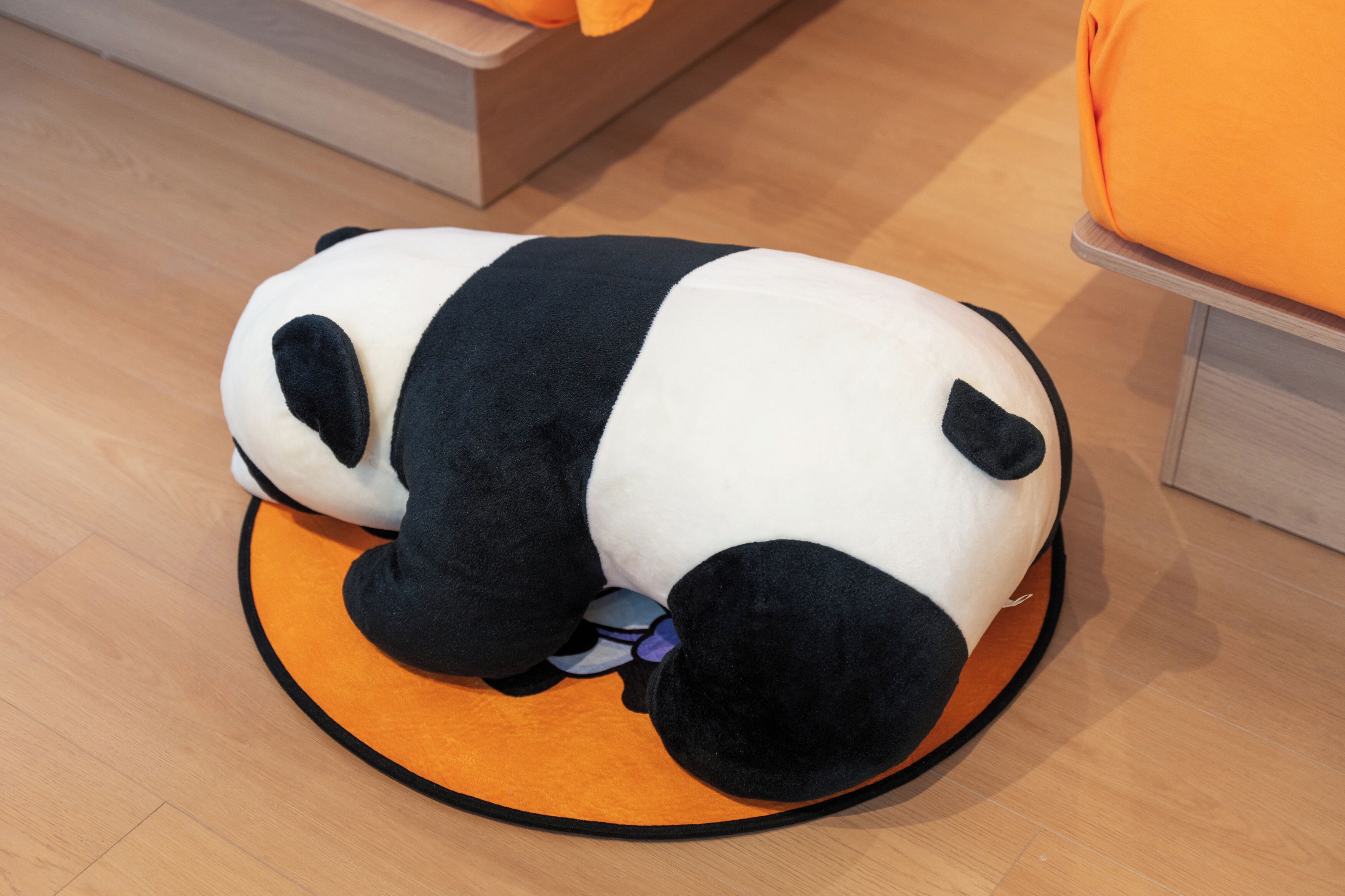 Um brinquedo de panda macio sobre um tapete laranja em um quarto com piso de madeira e parede em tom claro.