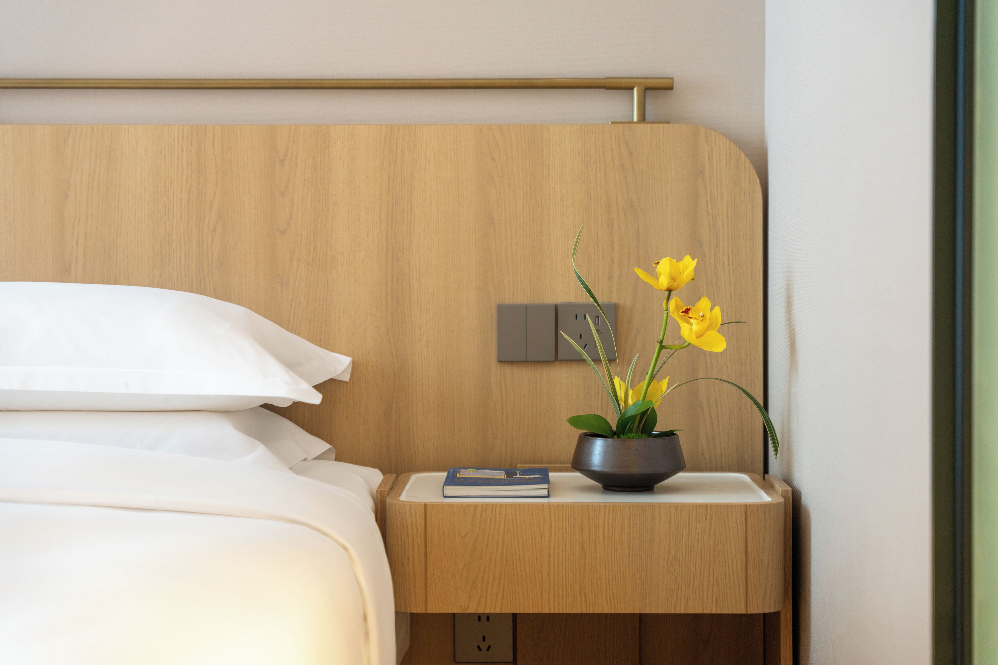 Una cama bien hecha con sábanas blancas, una mesita de noche de madera con un libro y una planta en maceta, y un interruptor de luz en la pared.