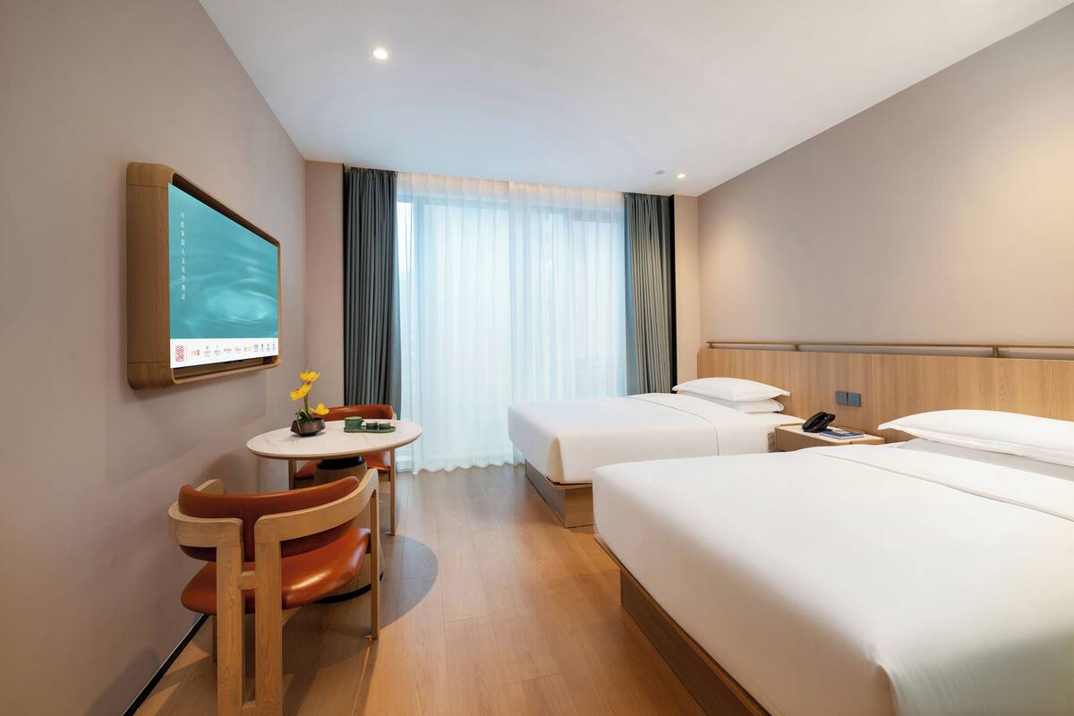 A hotel room with two beds, a desk with a chair, a TV mounted on the wall, and a large window with curtains.