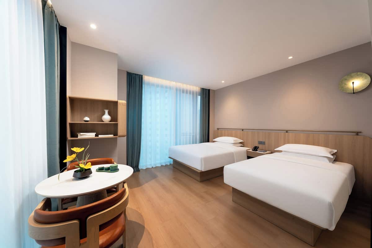 A hotel room with two beds, a small table with chairs, a vase with flowers, and a large window with curtains.