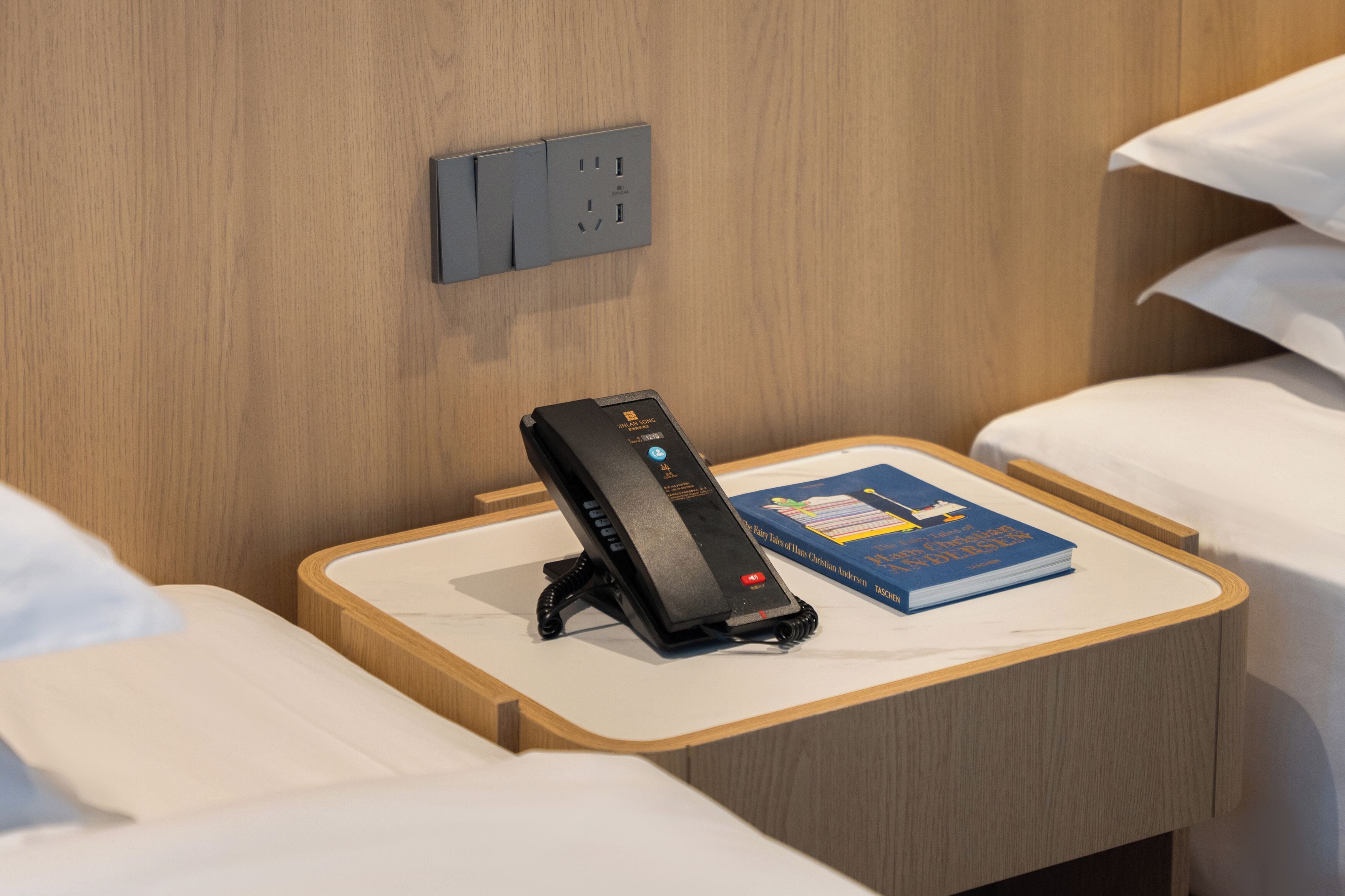 Een hotelkamer met nachtkastje, een boek, telefoon en een stopcontact.