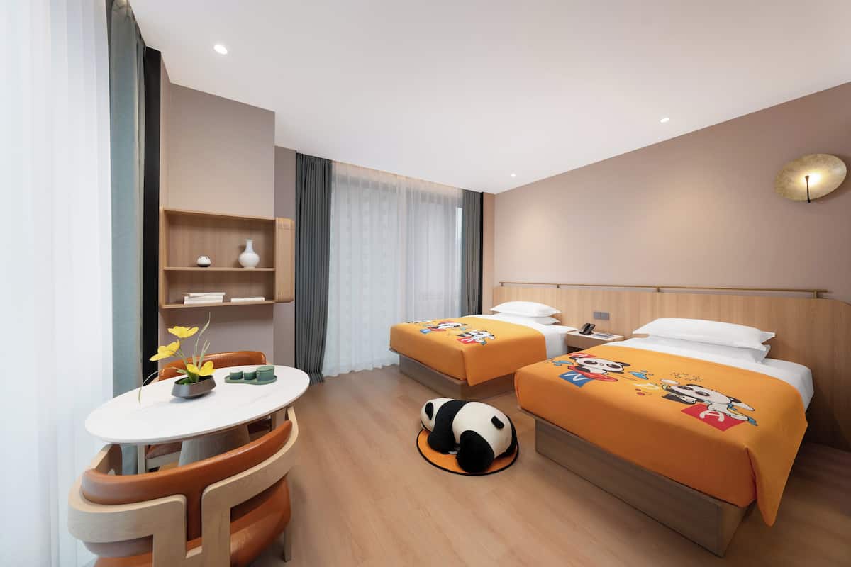 A hotel room with two beds, a small table, and a chair. There is a panda plush toy on the floor.