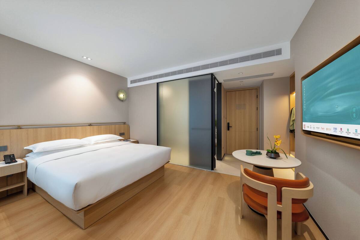 A modern hotel room with a large bed, a desk with a chair, a television, and a sliding door leading to another room.