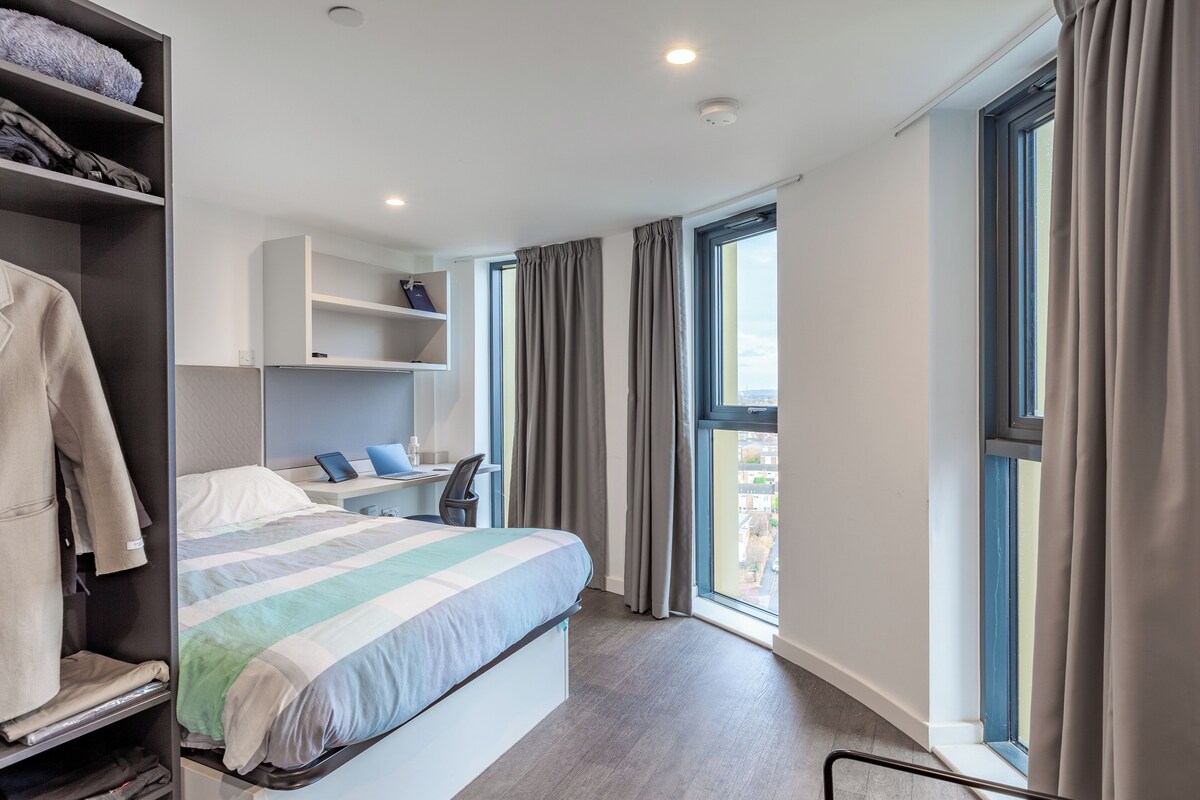 Ein modernes Hotelzimmer mit Bett, Schreibtisch, Kleiderschrank und großen Fenstern.