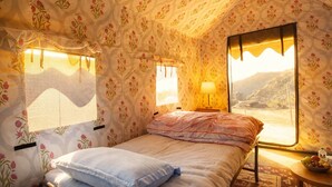 A cozy room with floral wallpaper, a bed with a patterned blanket, a bedside lamp, and a window with a view of a mountainous landscape. - Yao Adventure Base Camp Bhatta Falls (Mussoorie)