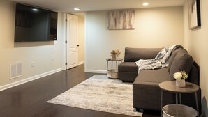 A spacious living room with a large flat-screen TV mounted on the wall, a grey sofa, a small round table with a vase, and a grey rug on the wooden floor. - Modern 4BR Severn Home • 10 Min to BWI • Near Fort Meade • Sleeps 8 (Severn)