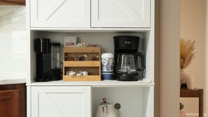 A white kitchen cabinet with two doors, a coffee maker, a kettle, and a shelf with kitchen items. - Modern 4BR Severn Home • 10 Min to BWI • Near Fort Meade • Sleeps 8 (Severn)