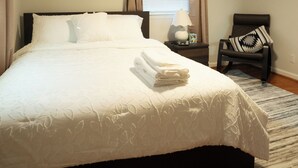A neatly made bed with a white comforter and pillows, a ceiling fan, a bedside lamp, a chair with a patterned pillow, and a window with curtains. - Modern 4BR Severn Home • 10 Min to BWI • Near Fort Meade • Sleeps 8 (Severn)