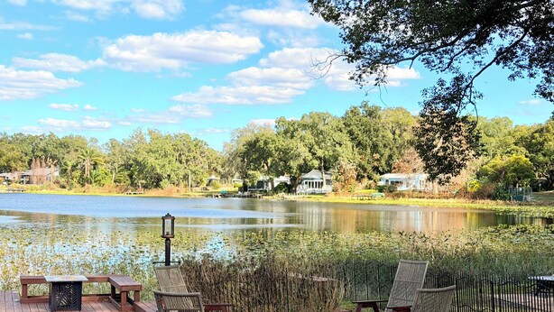 Une terrasse au bord du lac, avec des chaises en osier et un foyer, entourée d’une plateforme en bois et d’un chemin en briques.