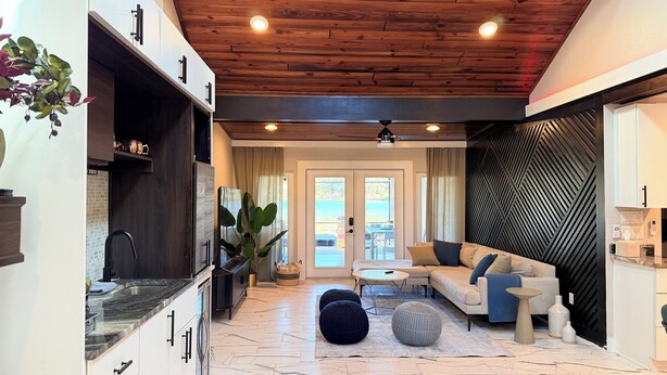 A modern kitchen with white cabinets and a black countertop, a living area with a beige sofa and round coffee table, and a wooden ceiling with recessed lighting.