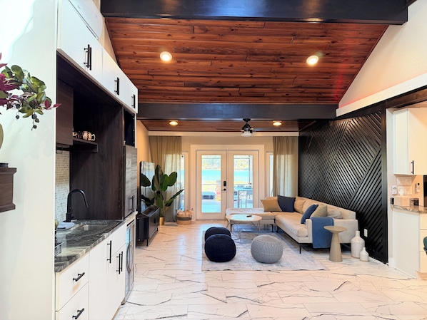 A modern kitchen with white cabinets and a black countertop, a living area with a beige sofa and round coffee table, and a wooden ceiling with recessed lighting.