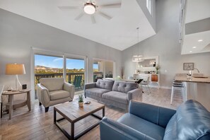 A spacious living area with a large window, a ceiling fan, and a kitchen area visible through the glass door. - IRB Intercoastal (Indian Shores)