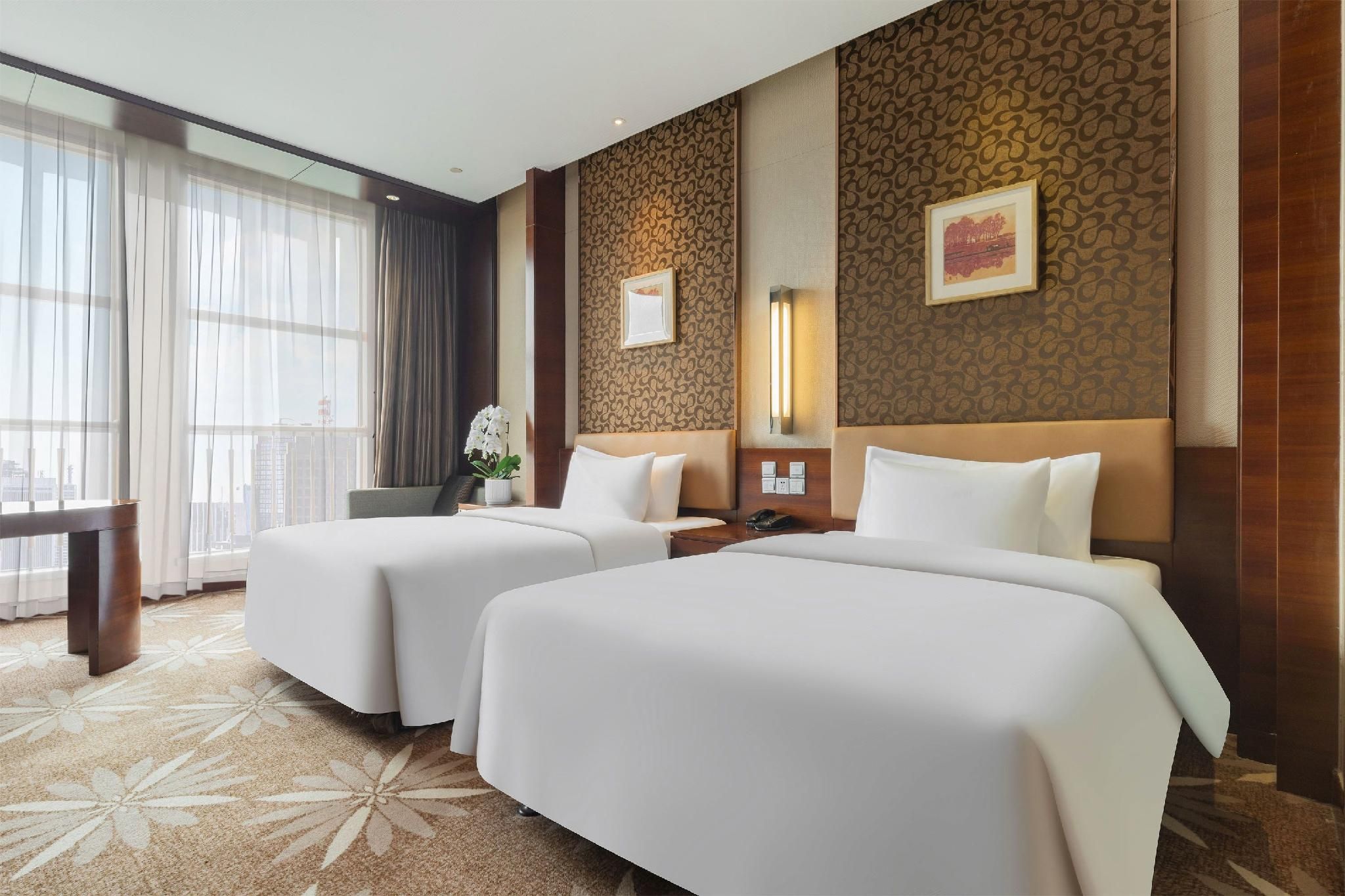 A hotel room with two beds, a desk, a chair, a potted plant, and a large window with curtains.
